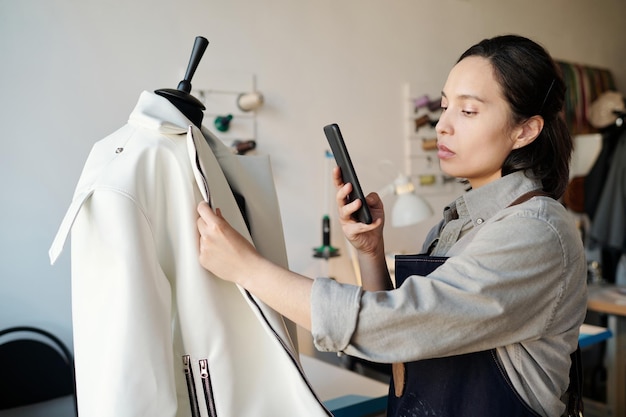 Jovem morena alfaiate em roupas de trabalho tirando foto de jaqueta em smartphone