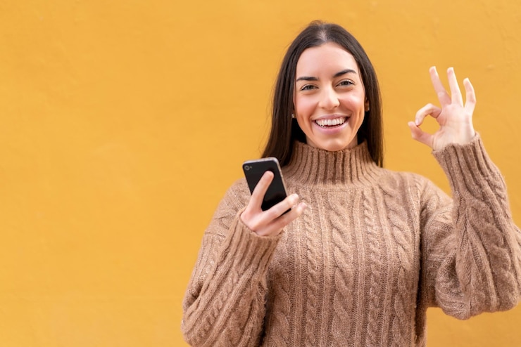 Foto jovem morena ao ar livre usando telefone celular e fazendo sinal de ok
