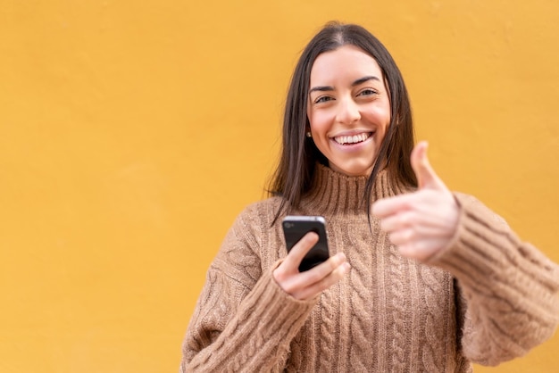 Jovem morena ao ar livre usando telefone celular enquanto faz polegares para cima