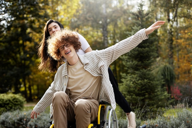 Jovem morena apóia seu amigo deficiente Jovem em uma cadeira de rodas feliz com a vida faz o gesto do avião ao ar livre