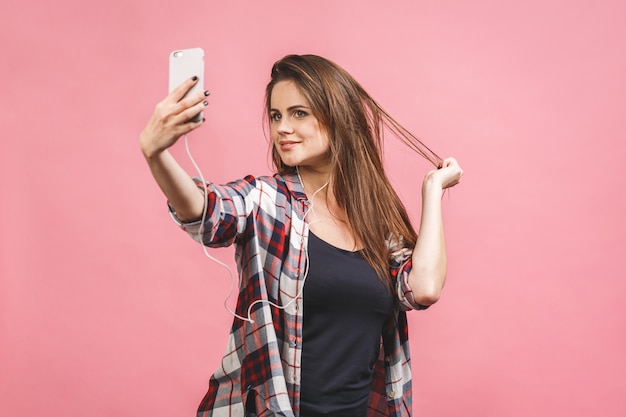 Jovem morena atraente alegre está sorrindo sobre o fundo rosa. Ela está tirando selfie na câmera do telefone, vestindo roupas casuais de verão.