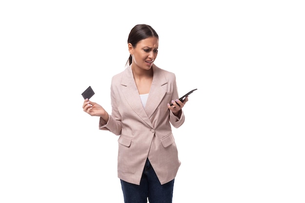 Jovem morena bem arrumada vestida com uma jaqueta bege usa um smartphone e segura um cartão de crédito
