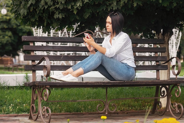 jovem morena bonita sentada ao ar livre no parque tomando café e olhando para o telefone