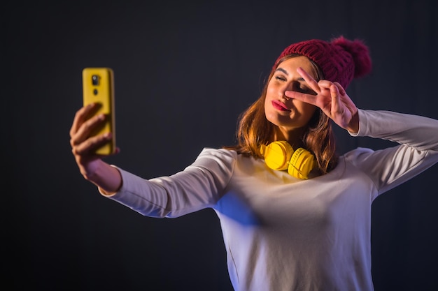 Jovem morena caucasiana com fones de ouvido amarelos, camiseta branca e chapéu de lã em um fundo preto, tirando uma selfie com o celular