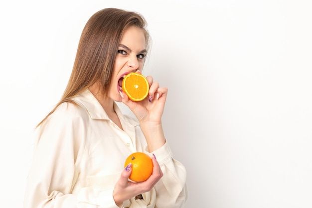 Jovem morena caucasiana muito astuta mordendo uma metade laranja e olhando para a câmera vestindo uma camisa branca sobre fundo branco