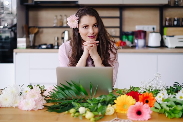 Jovem morena com cabelos longos ondulados trabalhando no laptop com flores na mesa com cozinha ao fundo florista talentosa desenvolvendo vendas on-line se preparando para a oficina