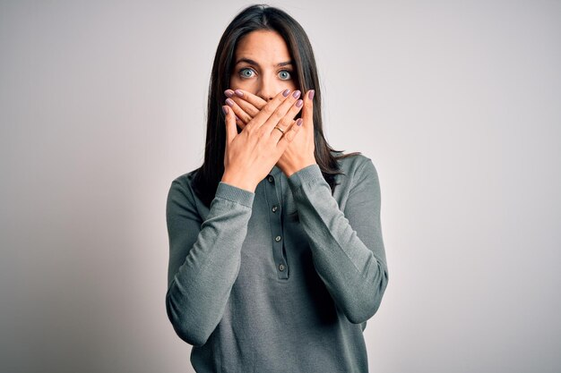 Foto jovem morena com olhos azuis, vestindo suéter verde casual sobre fundo branco chocado cobrindo a boca com as mãos por erro conceito secreto