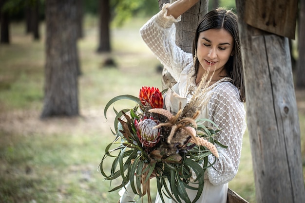 Jovem morena com um vestido branco com um buquê de flores na floresta em um fundo desfocado, copie o espaço.