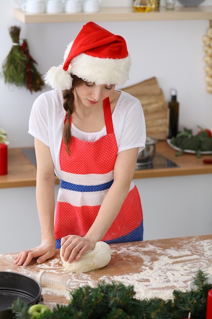 Jovem morena cozinhando pizza ou macarrão artesanal enquanto usava boné de Papai Noel na cozinha. Dona de casa preparando massa na mesa de madeira. Feliz Ano Novo, Casar com o Natal, Dieta, alimentação e saúde