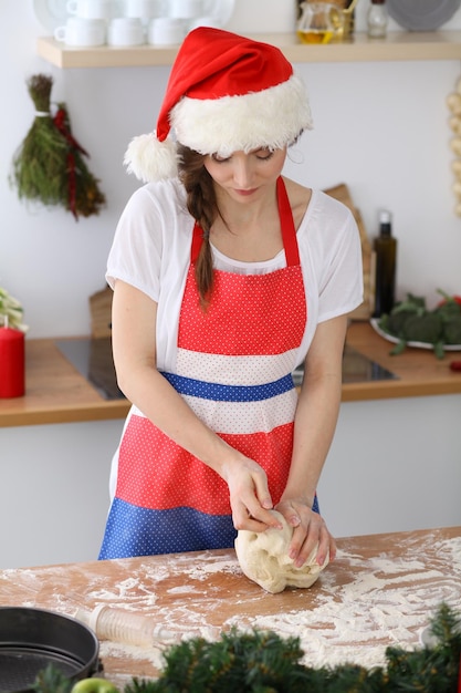 Jovem morena cozinhando pizza ou macarrão artesanal enquanto usava boné de Papai Noel na cozinha. Dona de casa preparando massa na mesa de madeira. Feliz Ano Novo, Casar com o Natal, Dieta, alimentação e saúde