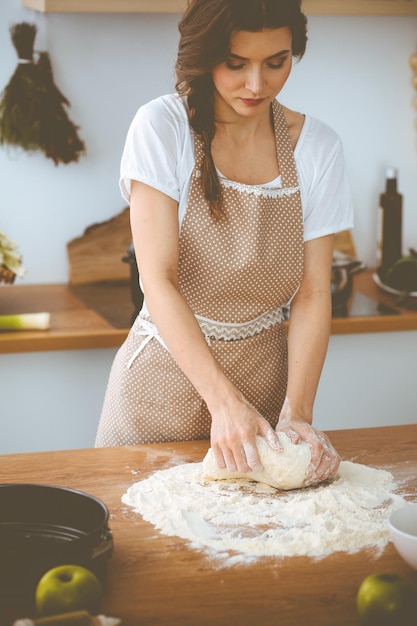 Jovem morena cozinhando pizza ou macarrão artesanal na cozinha. Dona de casa preparando massa na mesa de madeira. Conceito de dieta, comida e saúde.