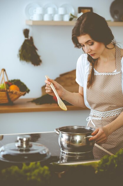 Jovem morena cozinhando sopa na cozinha. Dona de casa segurando a colher de pau na mão. Conceito de comida e saúde.