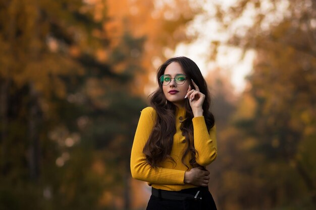 Jovem morena de cabelos compridos em jumper de malha amarela e óculos olhando para a câmera em pé contra a folhagem turva de árvores de outono no parque