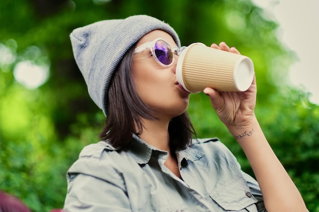 Jovem morena de chapéu bebe café de um copo de papel em um parque verde de verão.