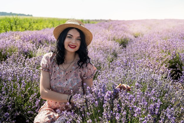 Jovem morena de chapéu de palha senta-se em um fundo de lavanda em um vestido de bolinhas brancas