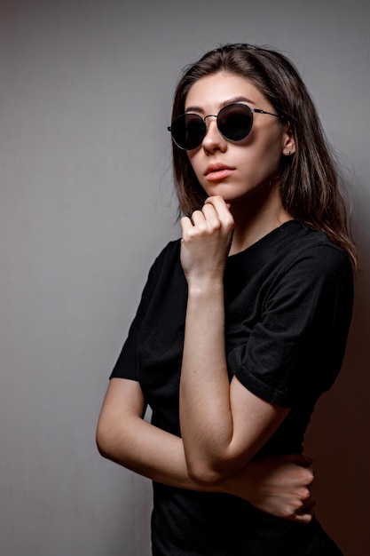 Foto jovem morena de óculos de sol pretos e uma camiseta preta no cinza