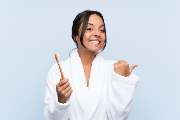 Jovem morena de roupão, escovando os dentes sobre fundo azul isolado, apontando para o lado para apresentar um produto