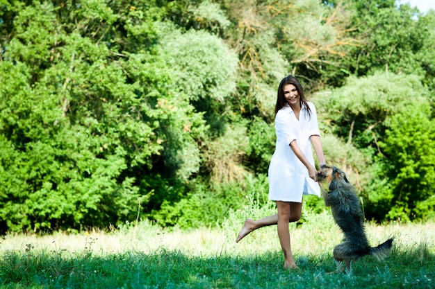 Jovem morena de vestido branco, brincando com cachorro na grama num dia de verão com árvores verdes e o rio