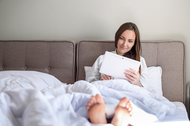 Jovem morena deitada na cama, usa um tablet moderno