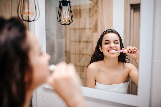 Jovem morena desportiva bonita fazendo rotina de manhã ou à noite no espelho. Ela limpa os dentes muito intensamente. Modelo olha no espelho. Corpo enrolado em toalha branca.