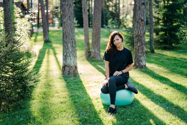 Jovem morena em forma descansa depois de fazer exercícios na bola de fitness leva poses de estilo de vida esportivo ativo em ambiente urbano na grama verde contra árvores com sol faz ginástica