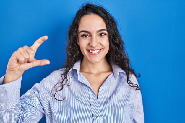 Foto jovem morena em pé sobre fundo azul sorrindo e confiante gesticulando com a mão fazendo sinal de tamanho pequeno com os dedos olhando e o conceito de medida de câmera