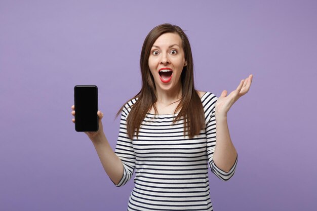 Jovem morena em roupas listradas casuais posando isolado no retrato de estúdio de fundo roxo violeta. Conceito de estilo de vida de pessoas. Mock up espaço de cópia. Segure o celular na mão com tela vazia em branco