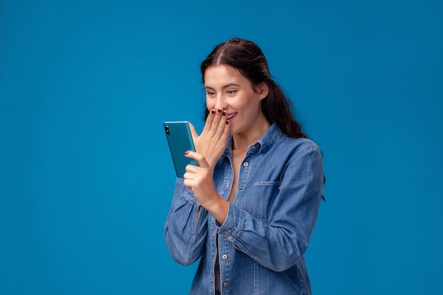 Jovem morena está posando com um smartphone de pé sobre fundo azul.