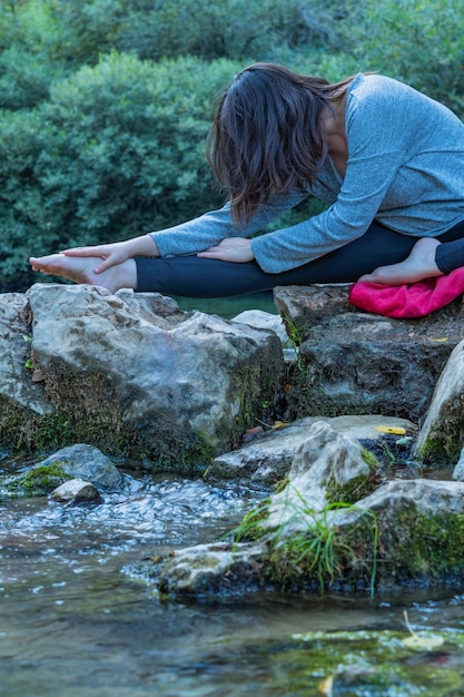 jovem morena fazendo ioga e meditação em um rio de alta montanha com um lago ao fundo. vestindo suéter azul e calça preta