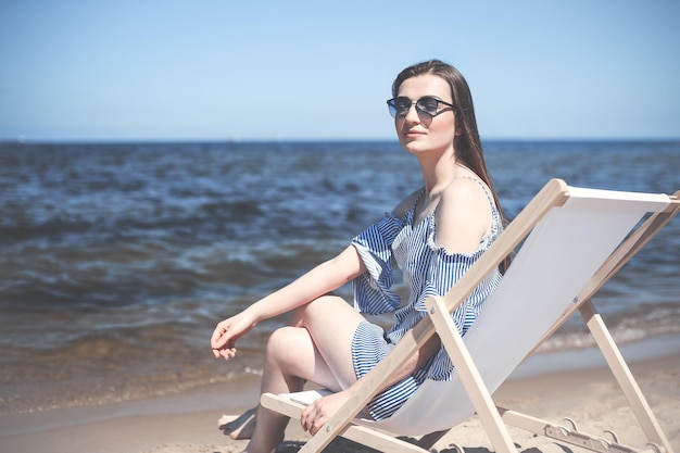 Jovem morena feliz relaxando em uma espreguiçadeira de madeira na praia oceânica enquanto olha para a câmera, sorrindo e usando óculos escuros da moda. O conceito de desfrutar de férias