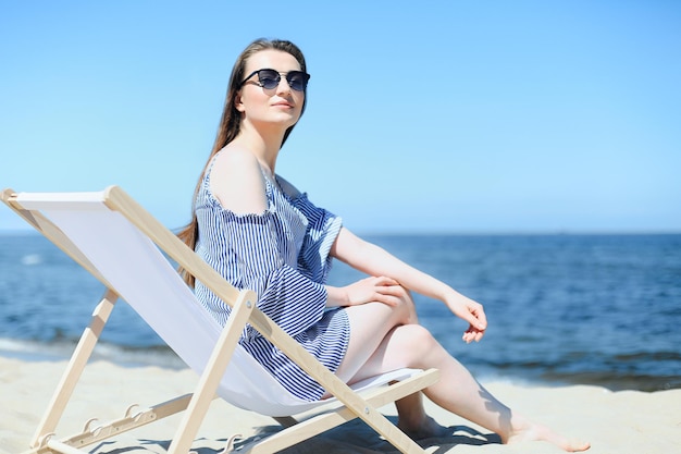 Jovem morena feliz relaxando em uma espreguiçadeira de madeira na praia oceânica enquanto sorri e usa óculos de sol da moda. O conceito de desfrutar de férias