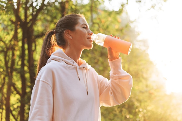 Jovem morena magra em roupas esportivas correndo e bebendo água na floresta na hora dourada do nascer do sol Saúde e bem-estar estilo de vida fitness