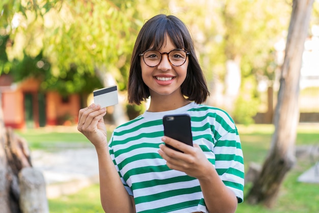 Jovem morena no parque comprando com o celular com cartão de crédito