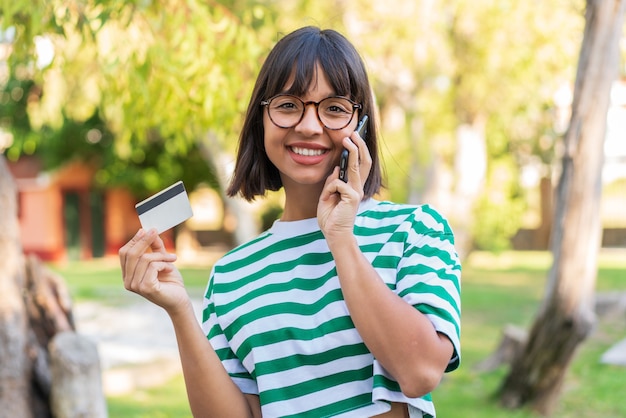 Jovem morena no parque conversando com o celular e segurando um cartão de crédito