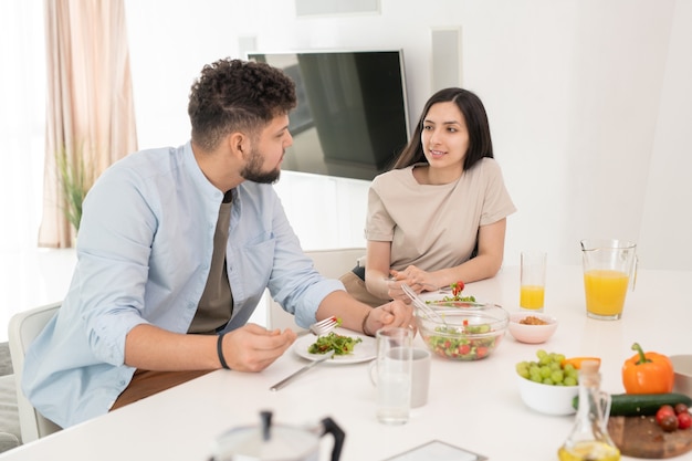 Jovem morena olhando para o marido durante uma conversa no café da manhã, enquanto os dois comem salada de legumes caseiros e suco de laranja