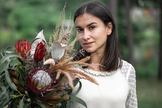 Jovem morena sedutora em um vestido branco com um buquê de flores na floresta em um fundo desfocado.