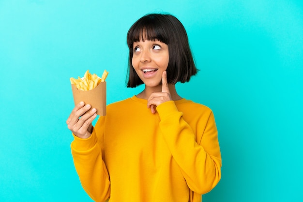 Jovem morena segurando batatas fritas sobre uma superfície azul isolada, pensando em uma ideia enquanto olha para cima