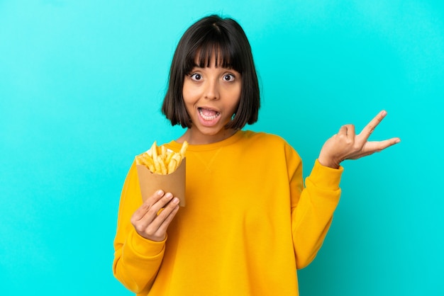 Jovem morena segurando chips fritos sobre uma superfície azul isolada com expressão facial chocada