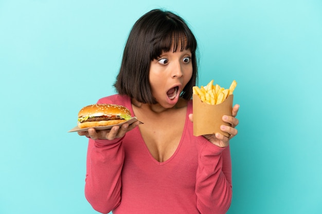 Jovem morena segurando hambúrguer e batatas fritas sobre fundo azul isolado