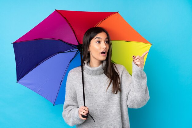 Jovem morena segurando um guarda-chuva sobre parede azul isolada, apontando para fora