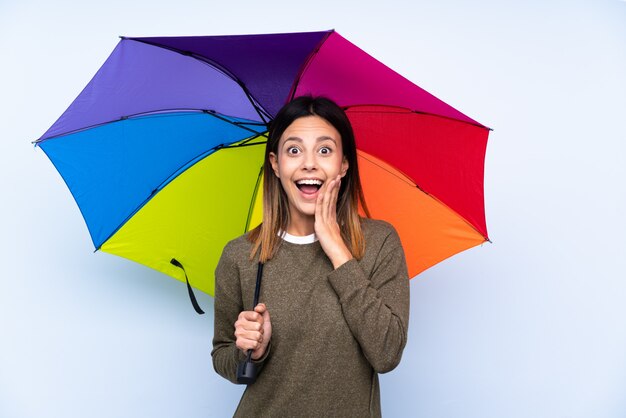 Jovem morena segurando um guarda-chuva sobre parede azul isolada com expressão facial de surpresa