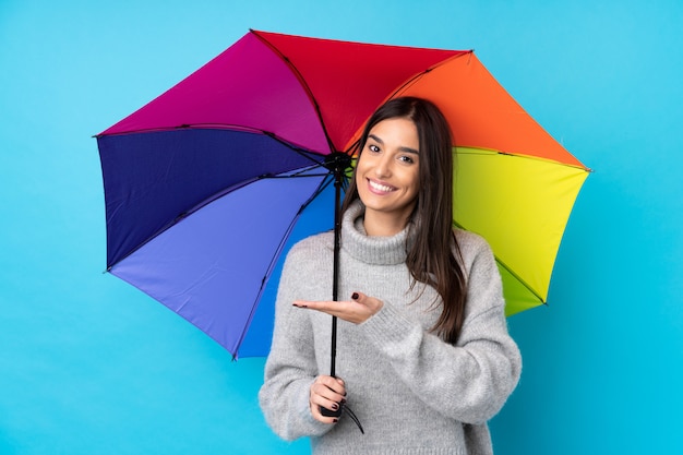 Jovem morena segurando um guarda-chuva sobre parede azul isolada, estendendo as mãos para o lado para convidar para vir