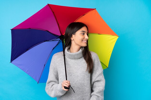 Jovem morena segurando um guarda-chuva sobre parede azul isolada, olhando para o lado