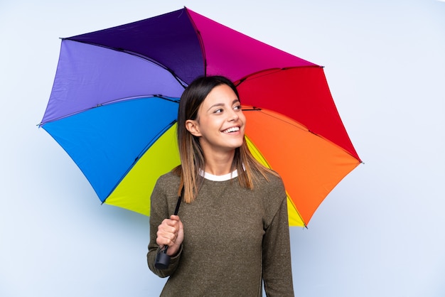 Jovem morena segurando um guarda-chuva sobre parede azul isolada, rindo e olhando para cima