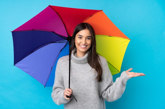 Jovem morena segurando um guarda-chuva sobre parede azul isolada, segurando copyspace imaginário na palma da mão