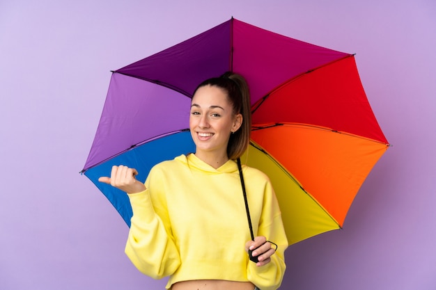 Jovem morena segurando um guarda-chuva sobre parede roxa isolada, apontando para o lado para apresentar um produto