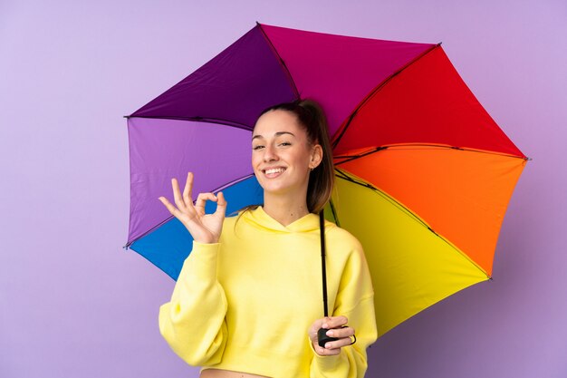 Jovem morena segurando um guarda-chuva sobre parede roxa isolada mostrando sinal de ok com os dedos