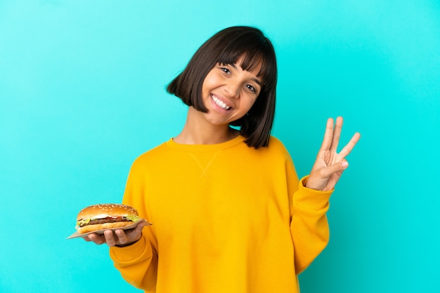 Jovem morena segurando um hambúrguer sobre um fundo isolado feliz e contando três com os dedos