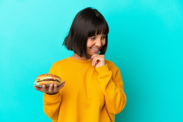 Foto jovem morena segurando um hambúrguer sobre um fundo isolado, olhando para o lado e sorrindo