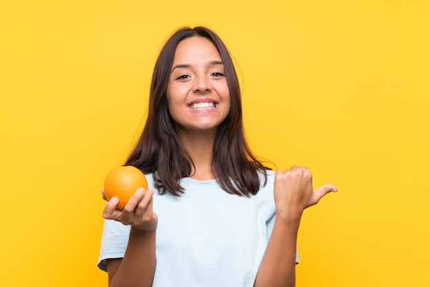 Jovem morena segurando uma laranja apontando para o lado para apresentar um produto